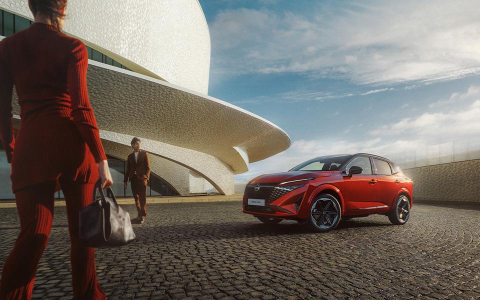 Ein roter Nissan Qashqai parkt auf einem gepflasterten Platz vor einem modernen Gebäude, zwei Personen in roter Kleidung gehen darauf zu.